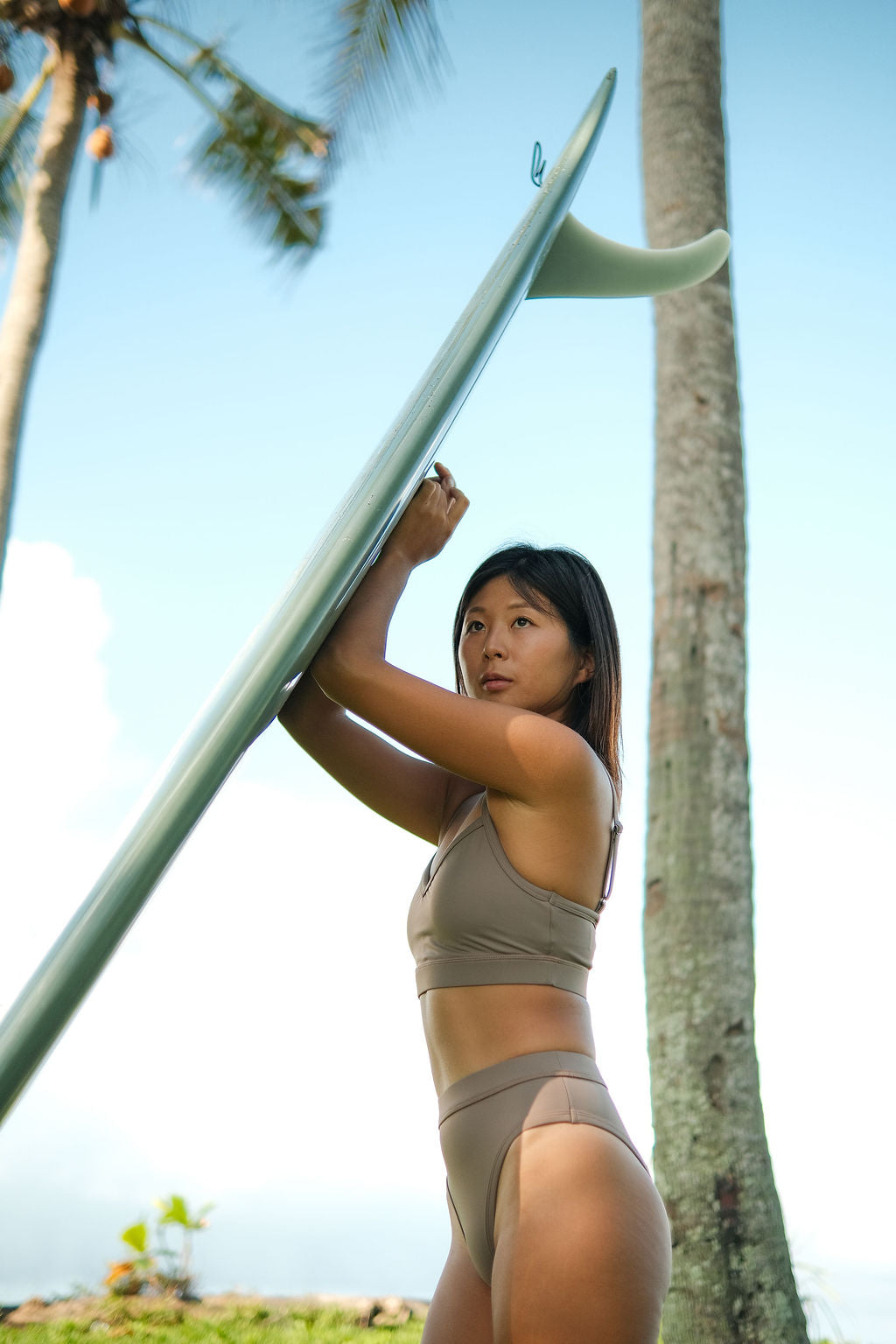Model wearing AFATO Rise Leg Bottom in mocha holding a surfboard – showcasing a natural surf lifestyle moment at the beach.